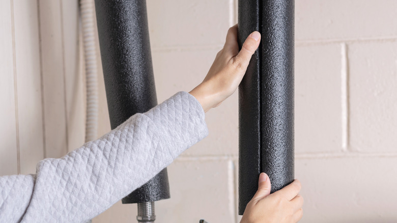A person in a sweater adding foam insulation tubes around water pipes in their home