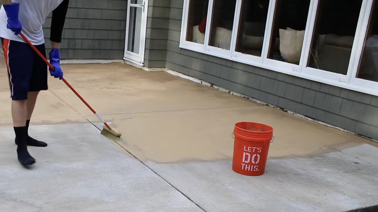 A person uses a broom to apply stain from an orange bucket to a concrete patio.