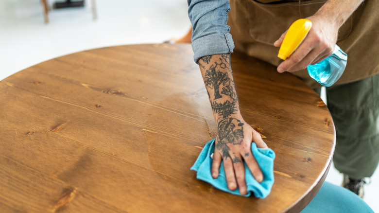 A person with tattooed hand cleans a wooden tabletop with a rag
