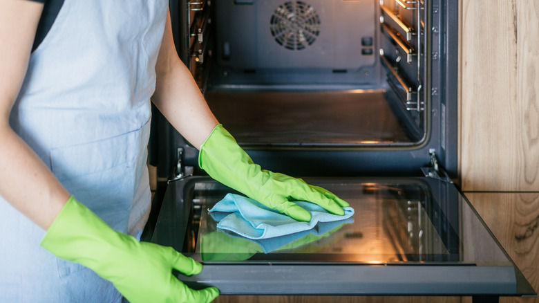 A woman wearing green gloves cleaning an oven door