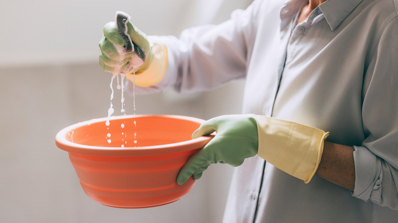 A person with gloved hands wringing out a cloth over a bowl of soapy water