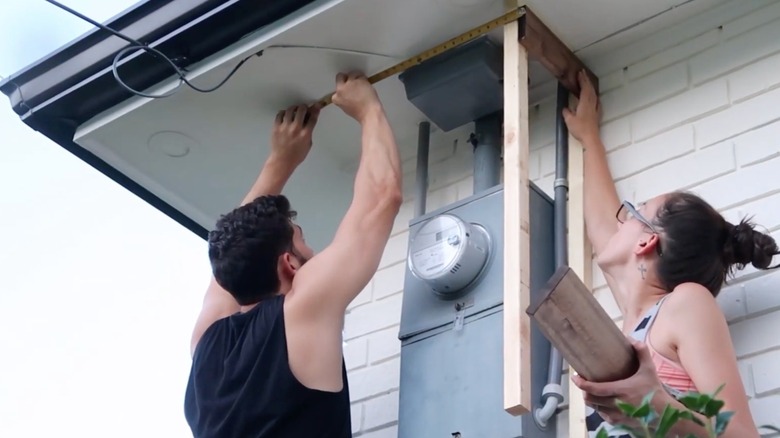 A couple install a wooden DIY cover cover a wall-mounted utility box on the side of their house.