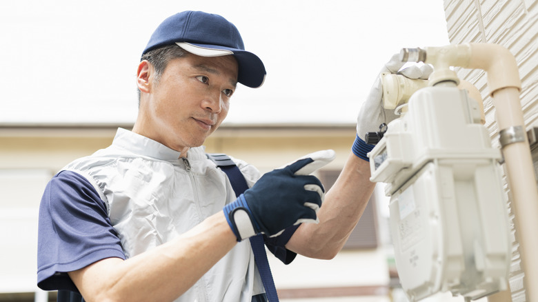 A service provider checks a wall-mounted gas meter on the side of a house.