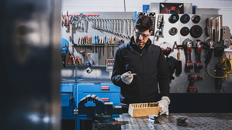 Man in a workshop examines his drill bits while wearing gloves and safety goggles