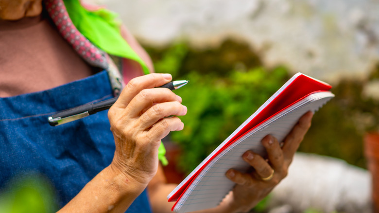 Person writing in a notebook with a pen in a garden.