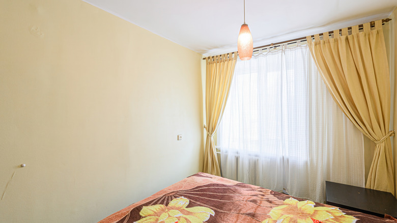 A room showing the side of a bed and an off-center window covered in yellow drapes pulled back with tiebacks