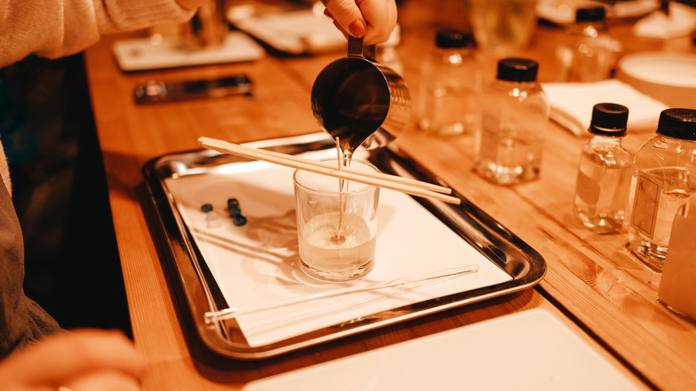 A person pours hot wax into a clear glass that has two wooden sticks holding up a wick