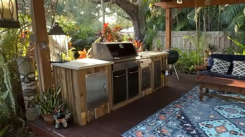 A wooden outdoor kitchen built around an existing grill with greenery in the backdrop on a wooden deck.