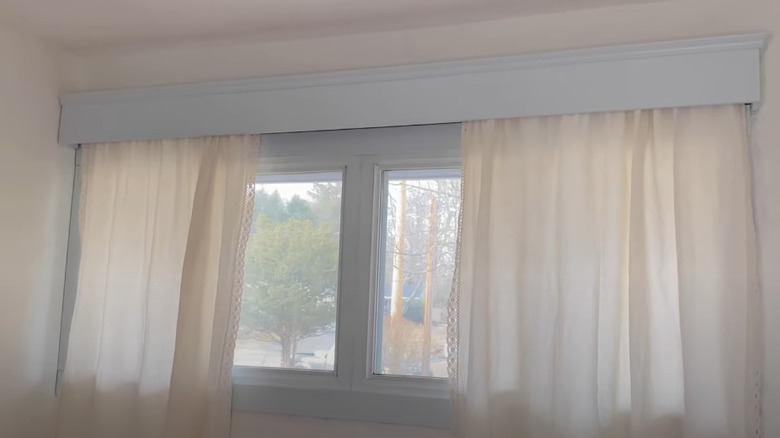 A white cornice with moldings at top over a beige, lace pair of curtains on a short window.