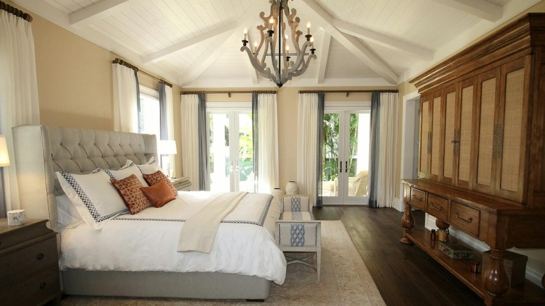 A large bedroom with trimmed window curtains, a large bed, statement chandelier, and armoire in the room.
