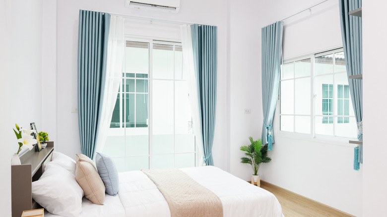 A bright white bedroom with a set of blue curtains hanging up and wide on each window.