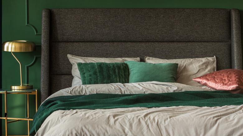 Emerald green walls with a bed with large brown headboard, beige bedding and green and pink cushions. There is a gold lamp on a gold bedside table.