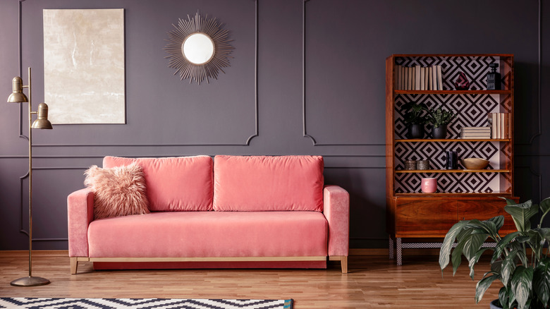 A living room with grey walls and a baby pink velvet sofa with fluffy cushion. There is a wooden bookcase with geometric backing and a golden freestanding lamp.