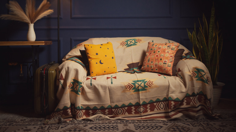 A living room with navy blue walls, side table with plant and a small sofa covered with a bohemian throw and cushions.