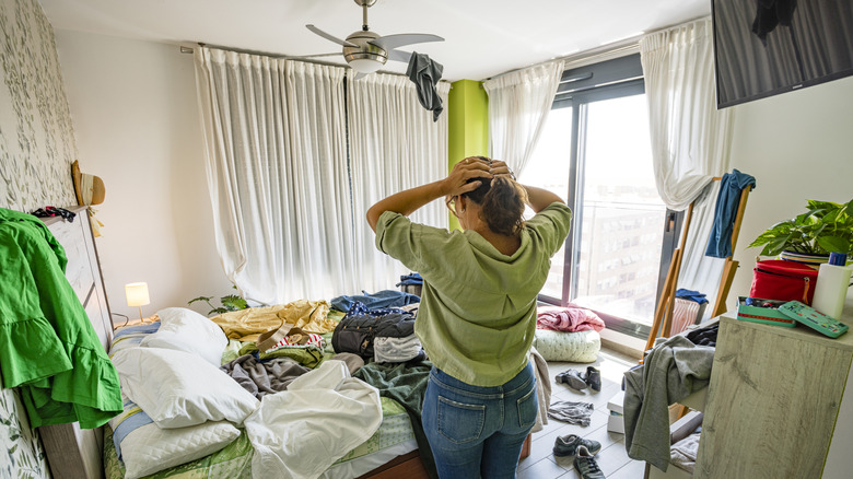 Woman standing in a cluttered bedroom with her hands on her head in despair