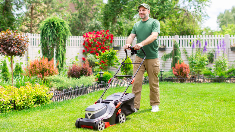A man mows his the lawn in his backyard next to a garden bed filled with flowering shrubs and small trees.