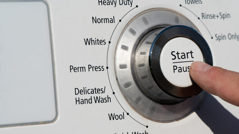 A finger presses the start-pause button on a washing machine with the dial set to delicate.