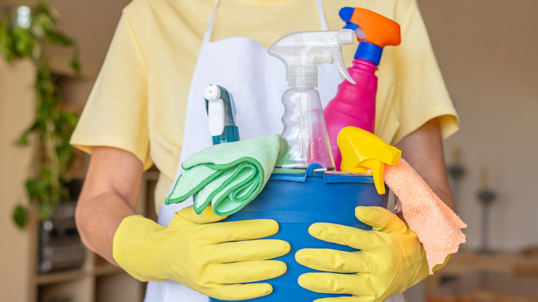 Person wearing yellow rubber gloves holding a collection of cleaning supplies in a blue bucket