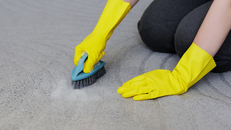 A person wearing yellow rubber gloves scrubbing a rug with a blue scrubbing brush