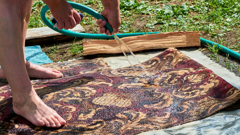 A person using a garden hose to rinse a rug outdoors