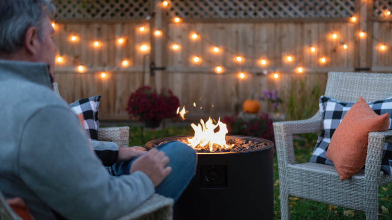 Man sitting in front of gas fire pit on a patio with a fence and lights in the background