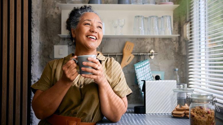 A healthy-looking older woman smiling in a kitchen setting while holding a mug