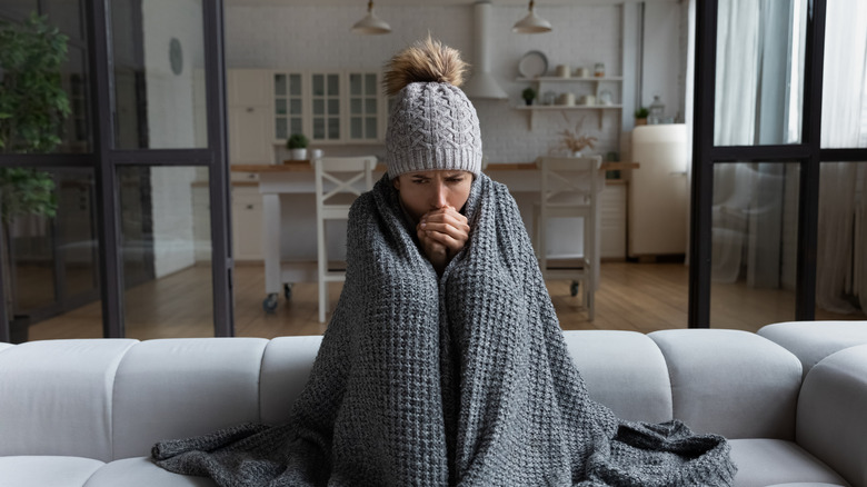 A cold woman wearing a hat sits shivering on a gray sofa