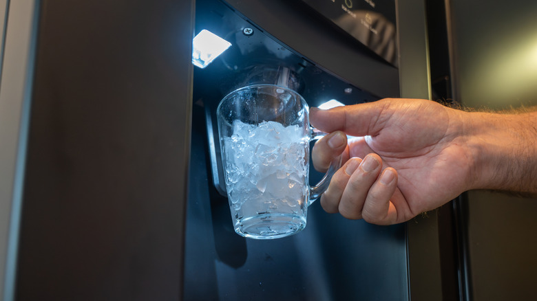 Hand holding a glass with ice from the ice dispenser in a fridge.