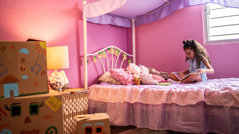 A little girl sitting on a pink princess bed with furry toys and reading a book.