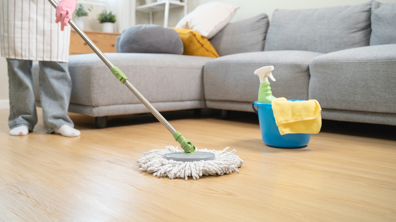 A person deep cleaning a laminate floor using a mop, blue bucket, and microfiber cloth with a grey couch in the background.