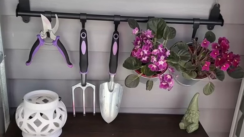 An IKEA HULTARP rail with garden tools and a couple of small potted flowering plants hanging from it.