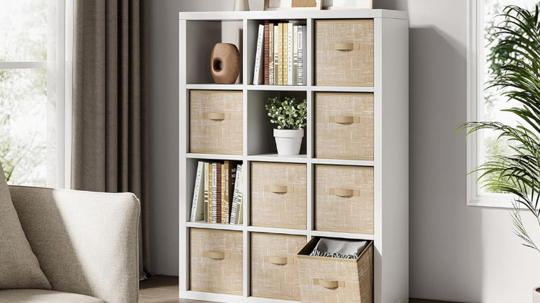 A white shelf with tan storage cubes, plants, books, and a vase in the cubbies.