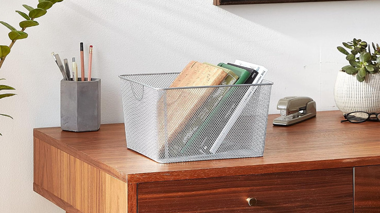 A wire mesh basket filled with books on top of a wood desk surrounded by office supplies
