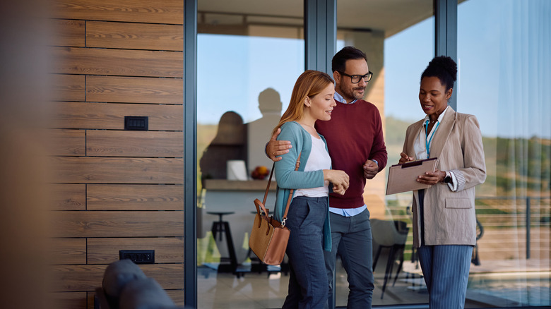 A real estate agent explains something on a clipboard to a couple in front of a home