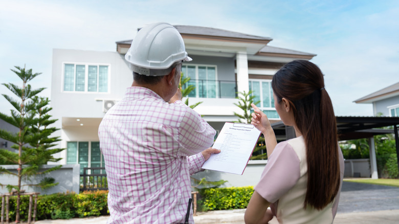 A male home inspector in a hard hat talks with a female homeowner outside of her home