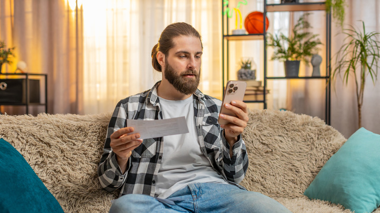 A man sits on couch holding his phone and utility bill