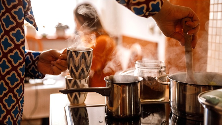 Person wearing a 1980s print holds a cup of steaming coffee while using a wooden spoon to stir a pot