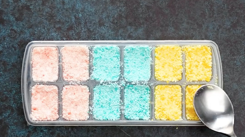 someone patting down with a spoon homemade bath bombs that are pink, blue, and yellow