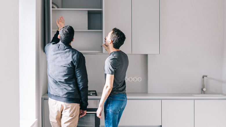 Two men discussing the design of a small, plain kitchen with boring white cabinetry.
