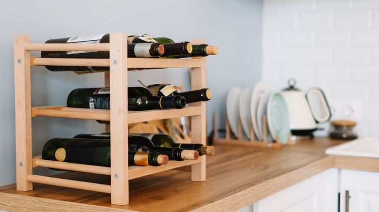 A wooden countertop with blurry dishes in the background and a small, wooden wine rack with wine bottles on it.