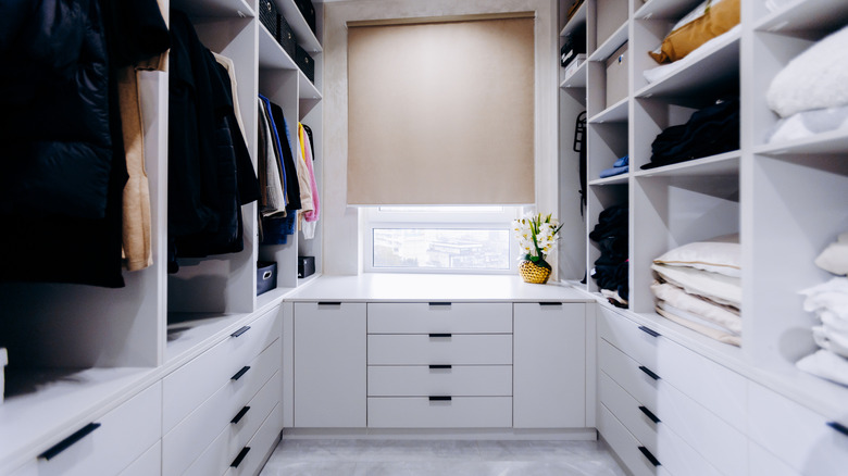 A white walk-in closet with hanging clothes on top and dresser drawers on the bottom with a sunny window at the back wall