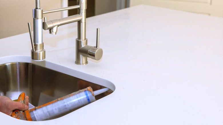 A person applies silicone sealant between a kitchen sink basin and quartz countertop