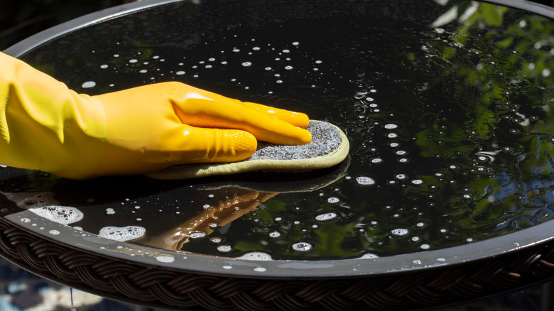 One hand in a yellow rubber glove gently cleans a glass table with a microfiber pad.