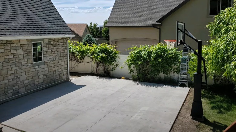 Stone house and concrete backyard with basketball hoop surrounded by lawn and shrubs