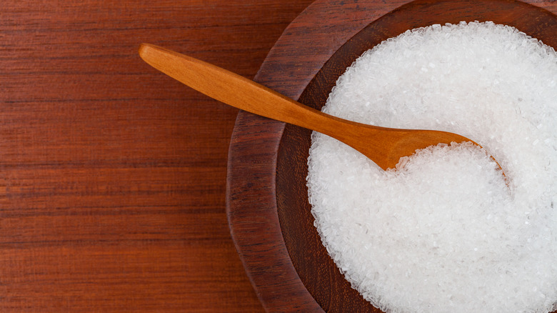 Epsom salt crystals are housed in a wooden bowl with a wooden spoon, sitting on a wood surface.