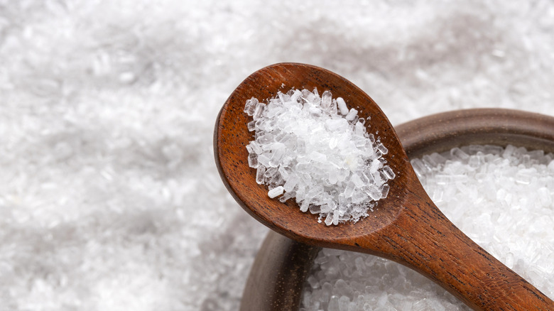 Epson salt in a wooden spoon on top of a wooden bowl full of white flakes.