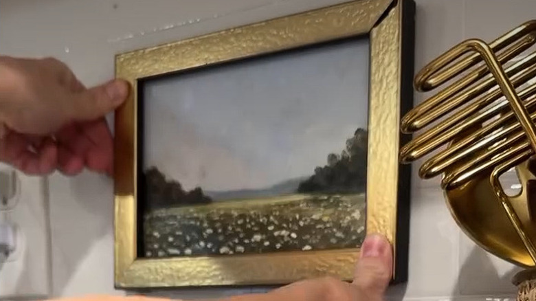A DIYer hot gluing a small, gold framed painting to a white tile wall.