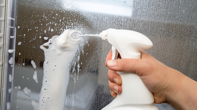 A person is spraying a foamy substance onto a glass shower door.