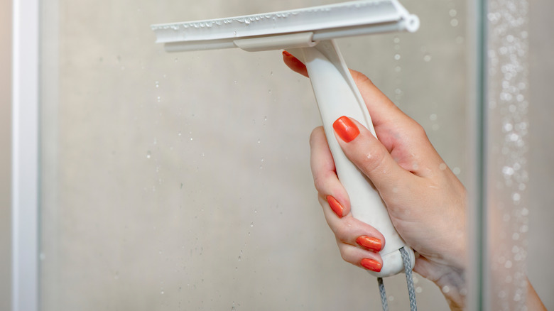 A female hand with orange-red nail polish holds a squeegee as she wipes down a shower door.