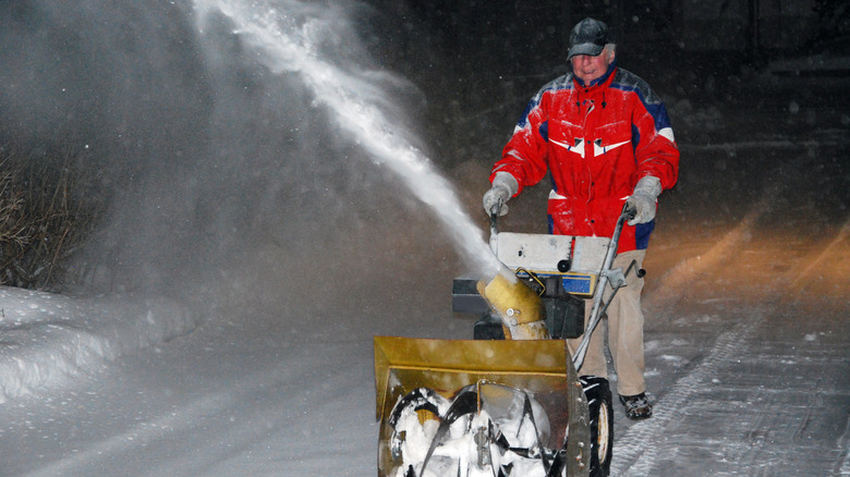 Man in red winter jacket uses snow blower at night.